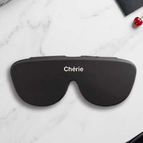 A pair of oversized black sunglasses with the word 'Chérie' printed in white across the lenses, arranged on a white marble surface with a camera, notebook, and small red heart as styled props.