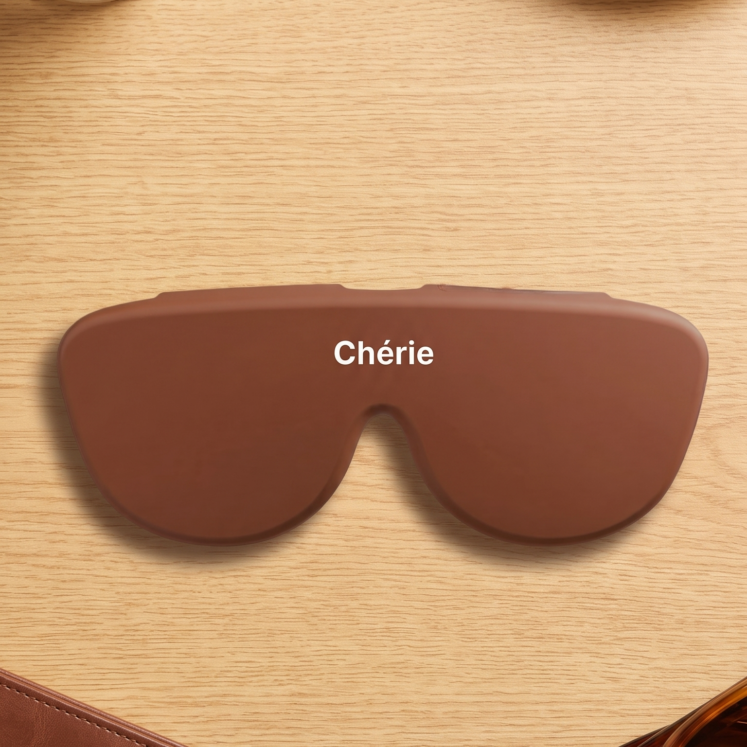 A brown heated eye mask with the name 'Chérie' printed in white text across the center, displayed on a light wood tabletop with lifestyle props including sunglasses, a wallet, an iced coffee, and a chocolate piece.