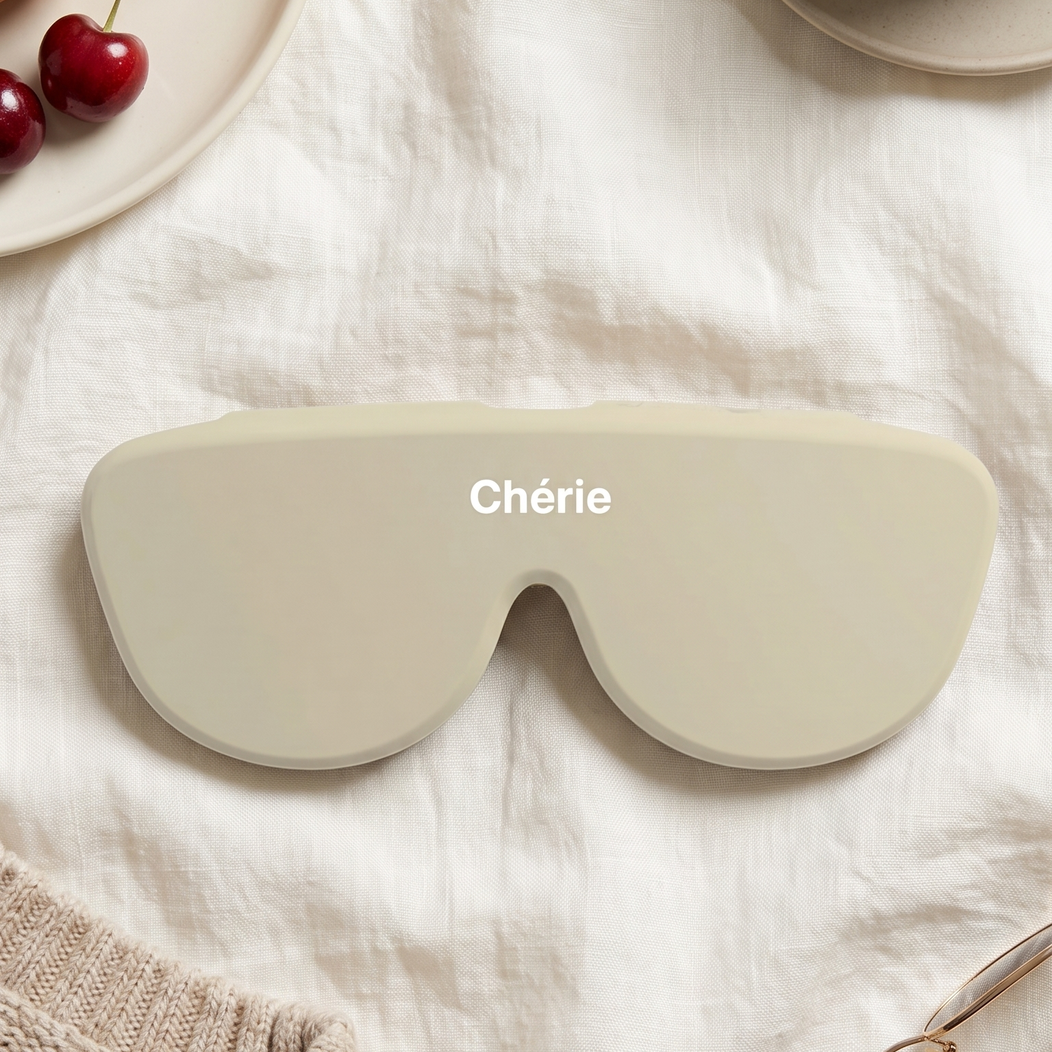 A beige sleep mask with the word 'Chérie' printed in white on the forehead, styled in a cozy flat-lay with a croissant, cherries, coffee, knit sweater, and eyeglasses on a white fabric background.