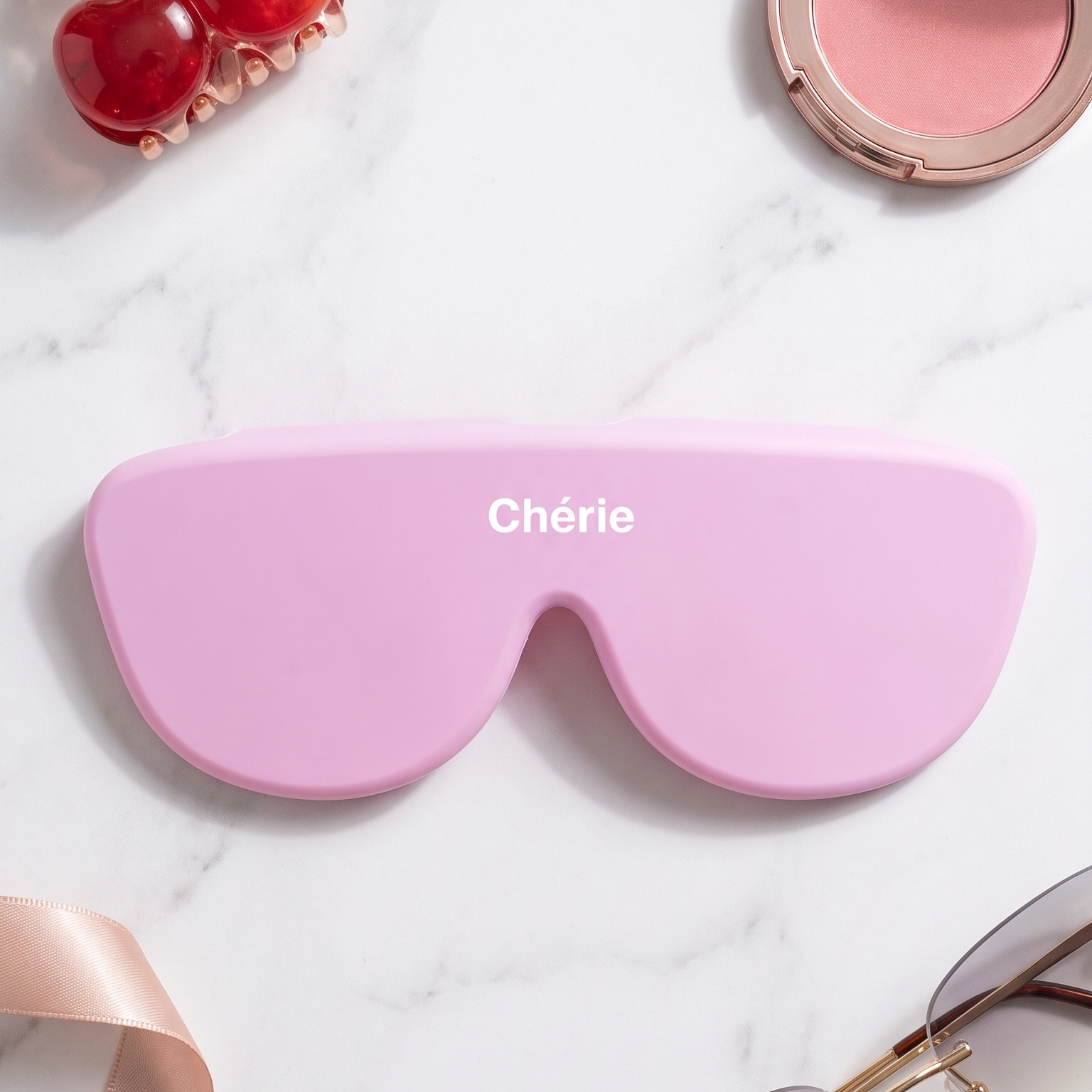 A pink aviator-style eyeglass case with the brand name 'Chérie' printed in white on the front, styled in a flat lay on a white marble background with cosmetics, sunglasses, a cherry-shaped hair clip, and a ribbon.