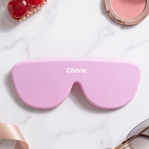 A pink aviator-style eyeglass case with the brand name 'Chérie' printed in white on the front, styled in a flat lay on a white marble background with cosmetics, sunglasses, a cherry-shaped hair clip, and a ribbon.