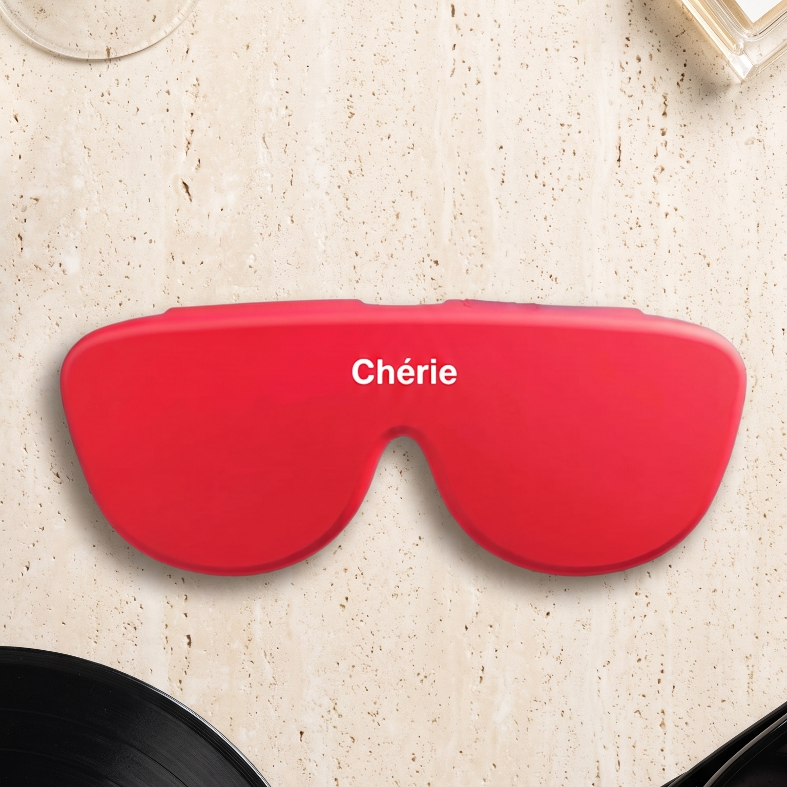 A bright red silicone sleep mask with the word 'Chérie' printed in white across the forehead, styled in a flat-lay arrangement on a light beige textured surface with a cocktail glass, perfume bottle, vinyl record, and sunglasses.